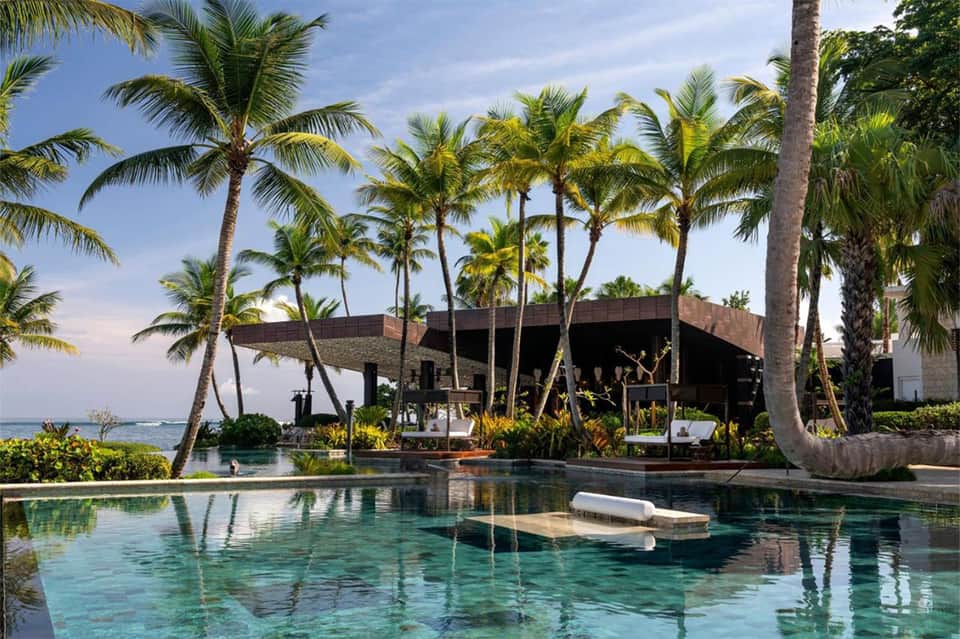 Luxury oceanfront pool with palm trees, modern dark building, and crystal-clear turquoise water at tropical resort