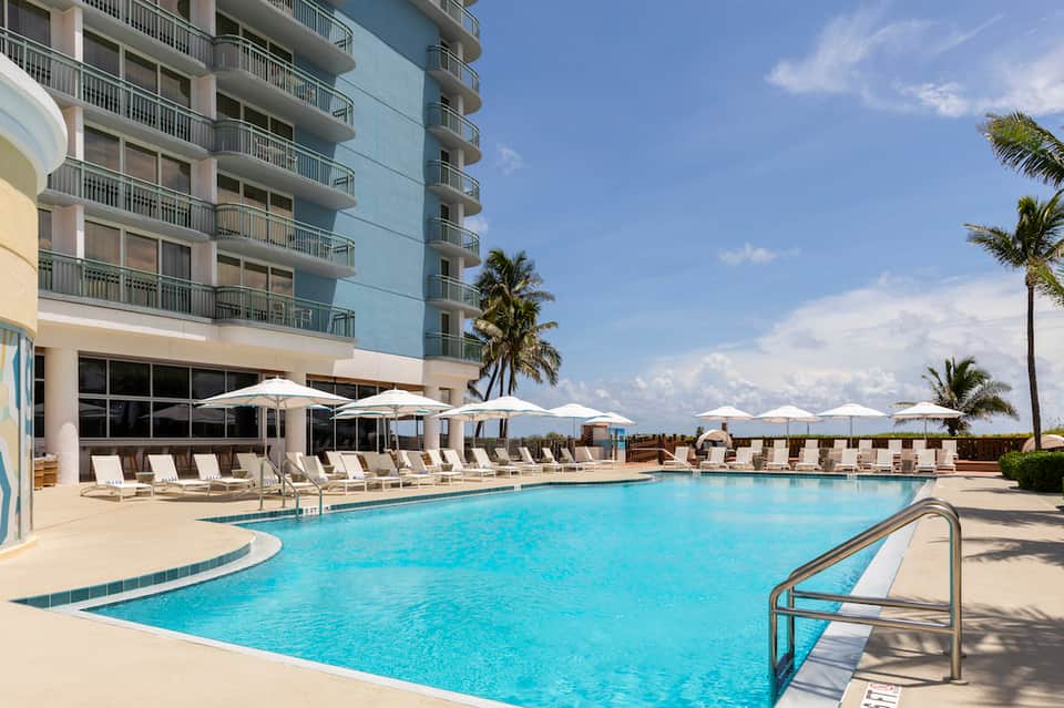 Resort infinity pool with lounge chairs and umbrellas, multi-story building with balconies, palm trees