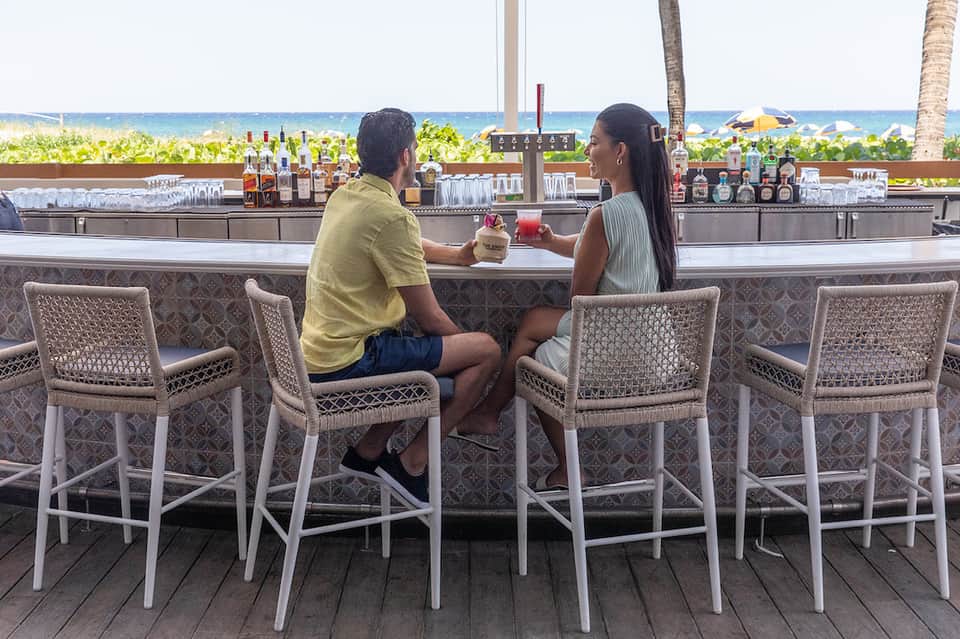 Couple enjoying cocktails at beachfront bar with ocean and palm trees in background