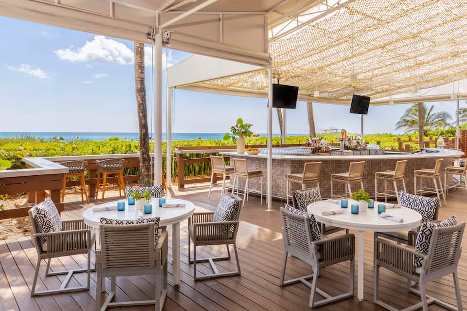 Beachfront bar and lounge with ocean views, woven shade structures, and white patio furniture on deck