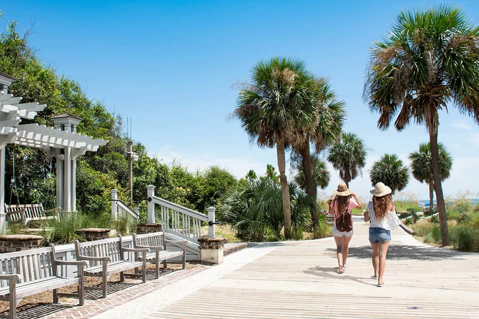 Wooden boardwalk with palm trees and beachgoers in wide-brimmed hats