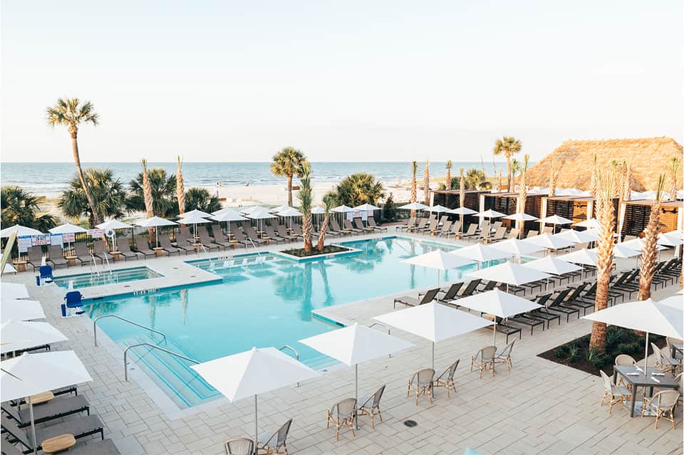 Large resort pool with multiple sections, palm trees, lounge chairs, and sandy beach with cliff backdrop