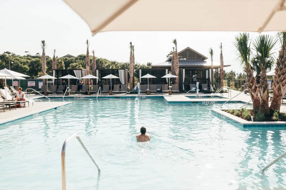Daytime resort pool with guests swimming, palm trees, white umbrellas, and poolside cabanas