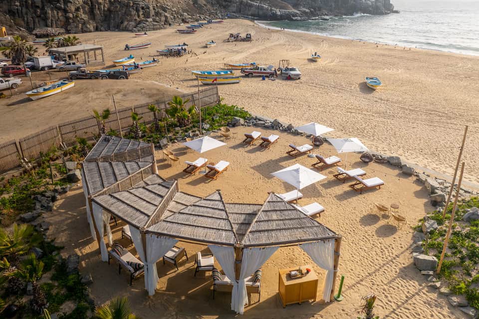 Beach scene with wooden cabanas, white umbrellas, boats, and rocky cliffs in background