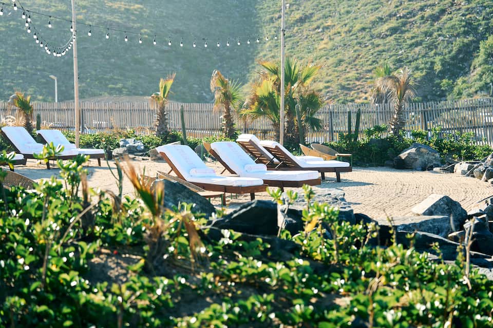 Beach lounge area with white cushioned sun loungers, palm trees, and mountainous backdrop