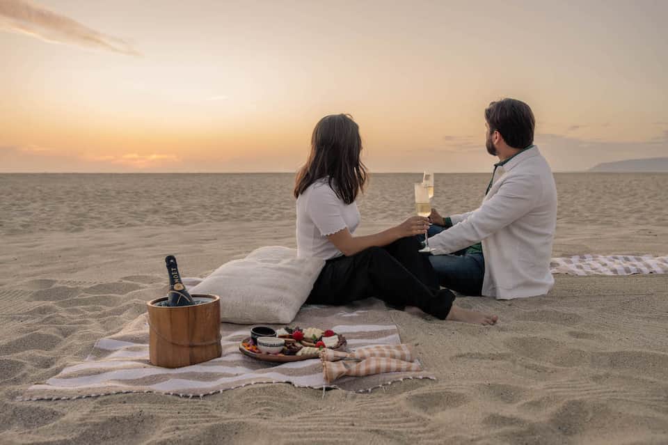 Couple enjoying champagne on beach at sunset with charcuterie board and ice bucket
