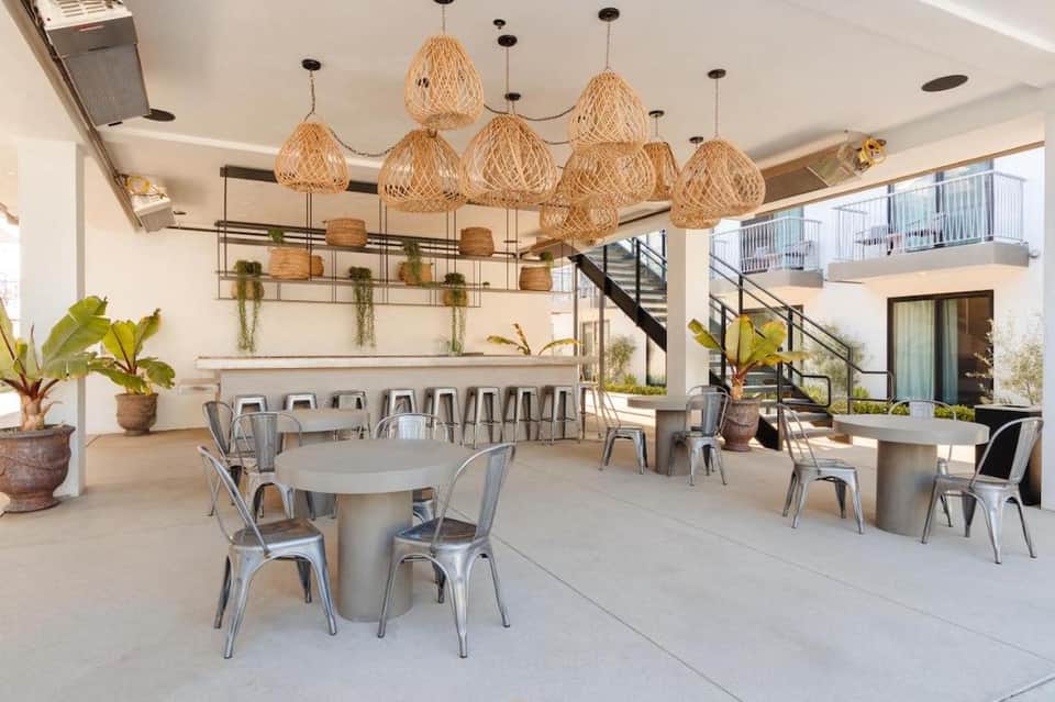 Modern hotel common area with woven pendant lights, round tables, metal chairs, potted plants, and a bar counter.