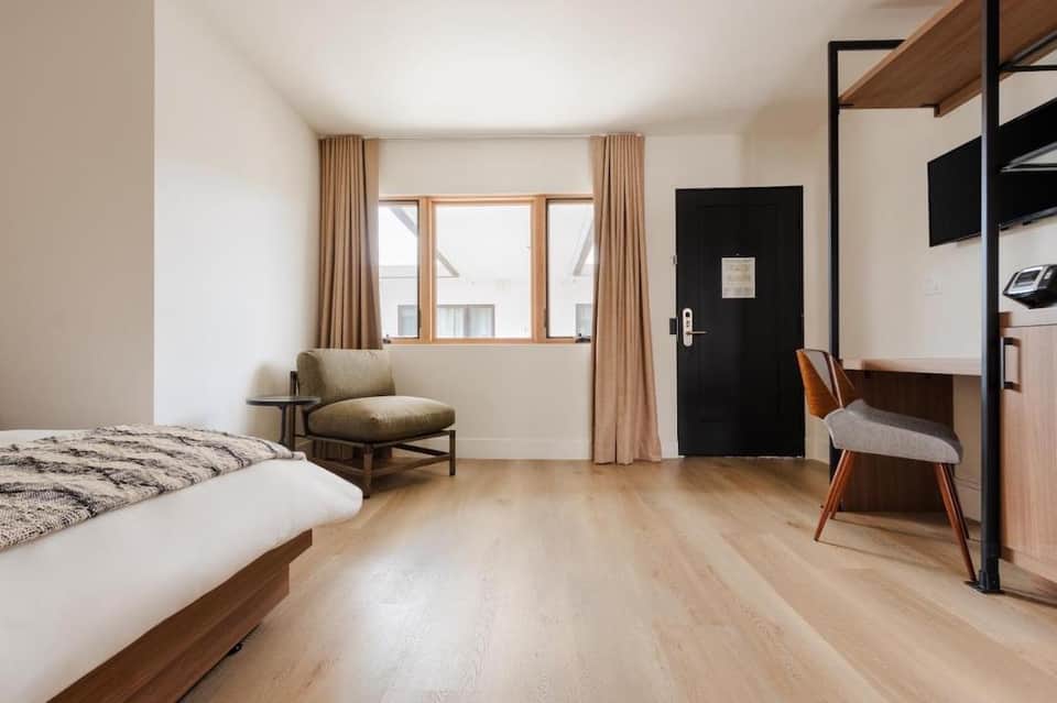 Minimalist hotel bedroom with wooden bed frame, tan chair, large window with curtains, and wood flooring