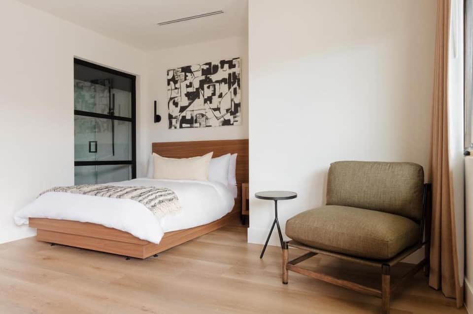 Modern hotel bedroom with wooden bed frame, earth-tone chair, and minimalist decor