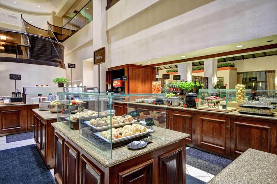Upscale hotel bakery with glass display cases showcasing pastries and baked goods, wood cabinetry, and multi-level seating area