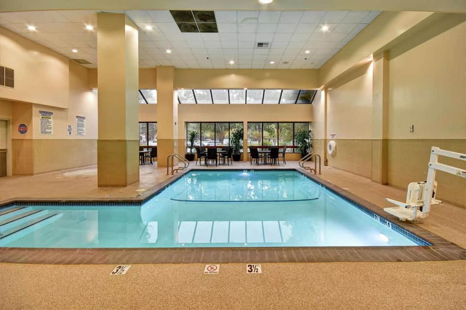 Indoor swimming pool with lane markers, beige walls, columns, and windows overlooking outdoor seating area