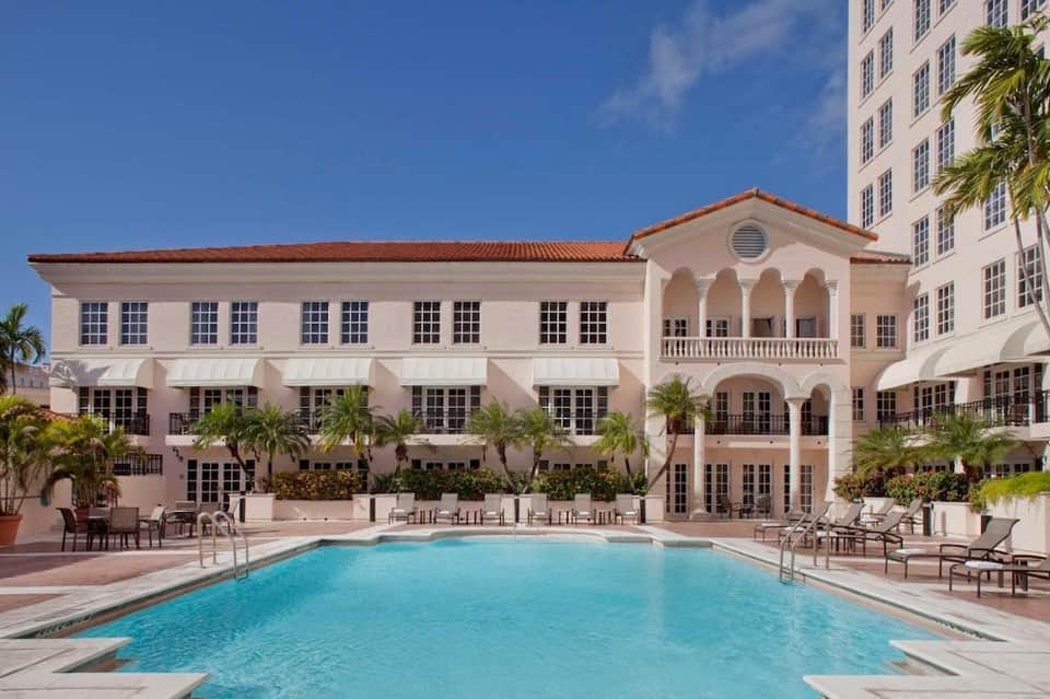 Pink neoclassical hotel building with pool, lounge chairs, and palm trees in courtyard setting
