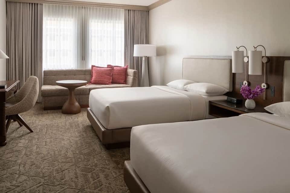Modern hotel room with two queen beds, upholstered headboard, sitting area with sofa, and floor-to-ceiling curtains