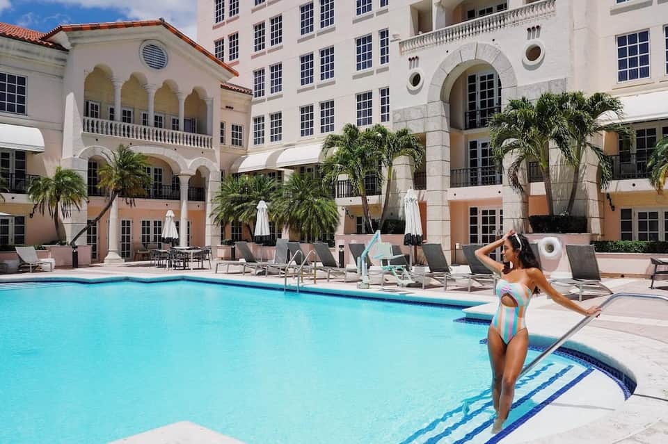 Elegant resort pool surrounded by classical pink and white buildings, palm trees, and umbrella-shaded lounge seating