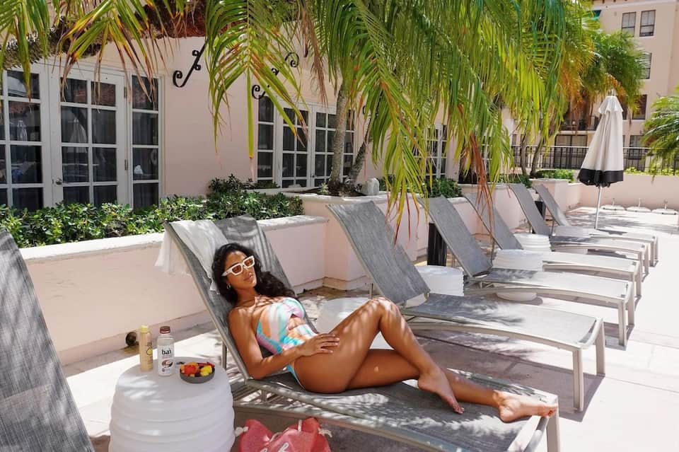 Woman relaxing on poolside lounge chair under palm trees at resort courtyard with white umbrellas