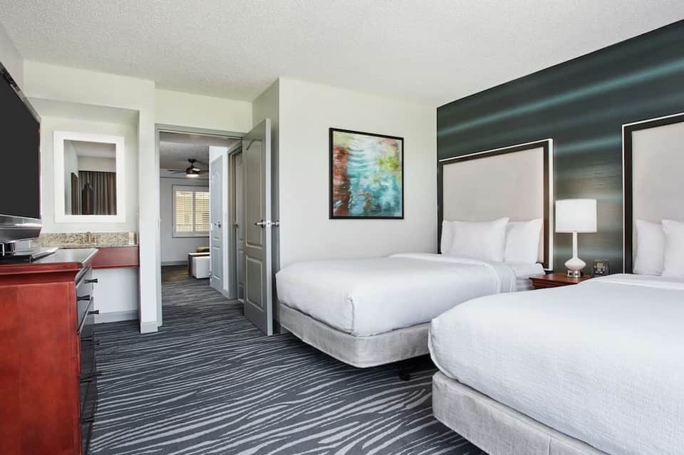 Double queen hotel room with striped bedding, red accent desk, and bathroom entrance
