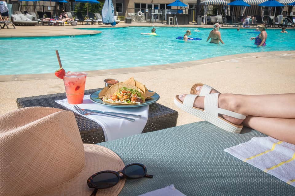 Poolside dining scene with colorful cocktail and plate of food, swimmers in turquoise pool background