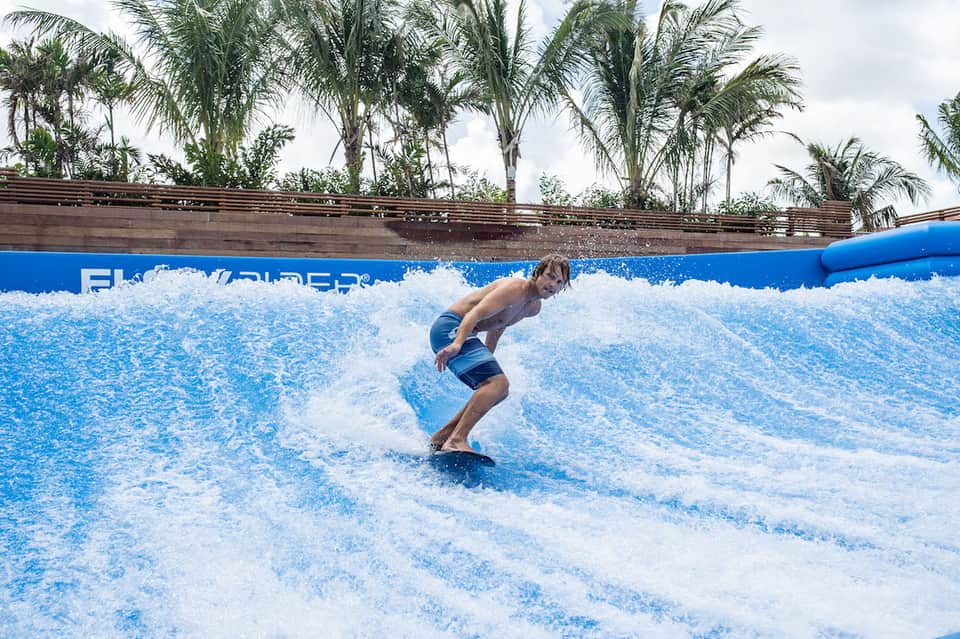 Man surfing on artificial wave at FlowRider water park surrounded by palm trees