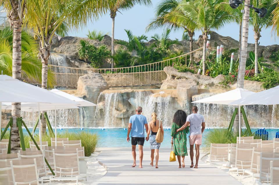 Resort pool area with waterfall feature, palm trees, white umbrellas, and rocky landscape backdrop