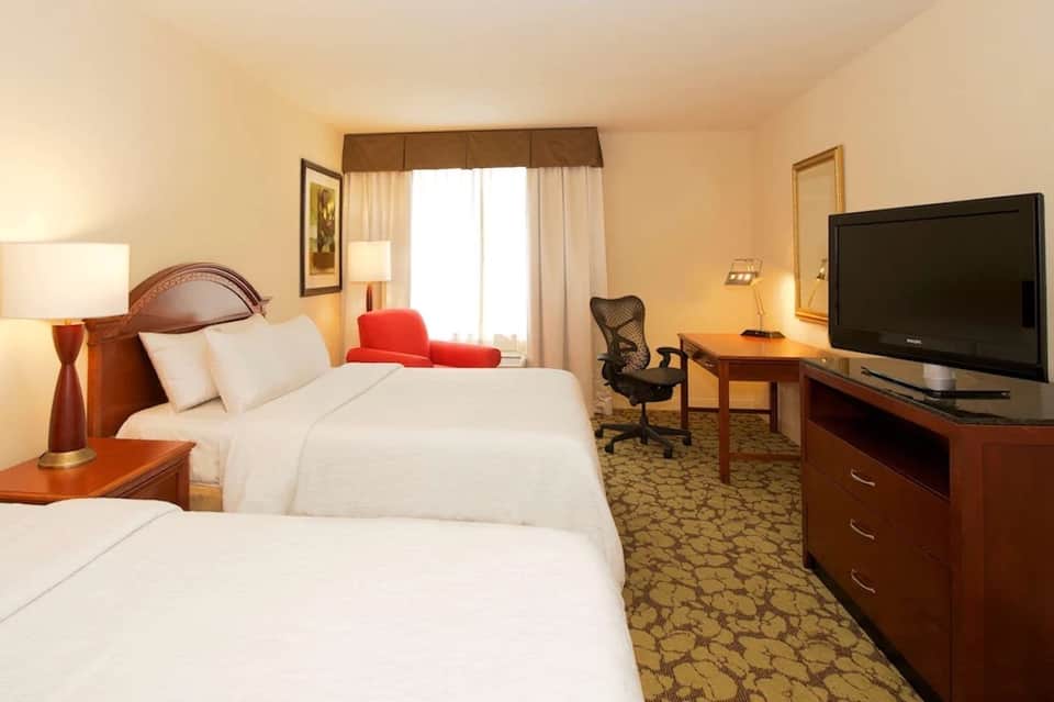 Hotel guest room with two queen beds, warm lighting, desk area, and red accent pillows by window