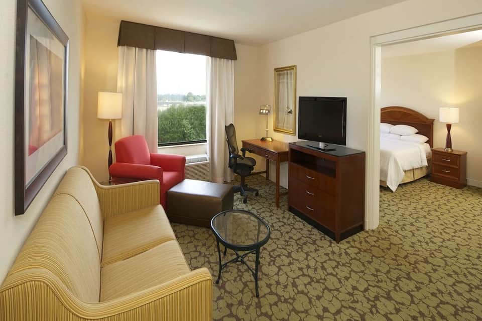 Hotel suite with living area, bedroom, desk, and window overlooking grounds