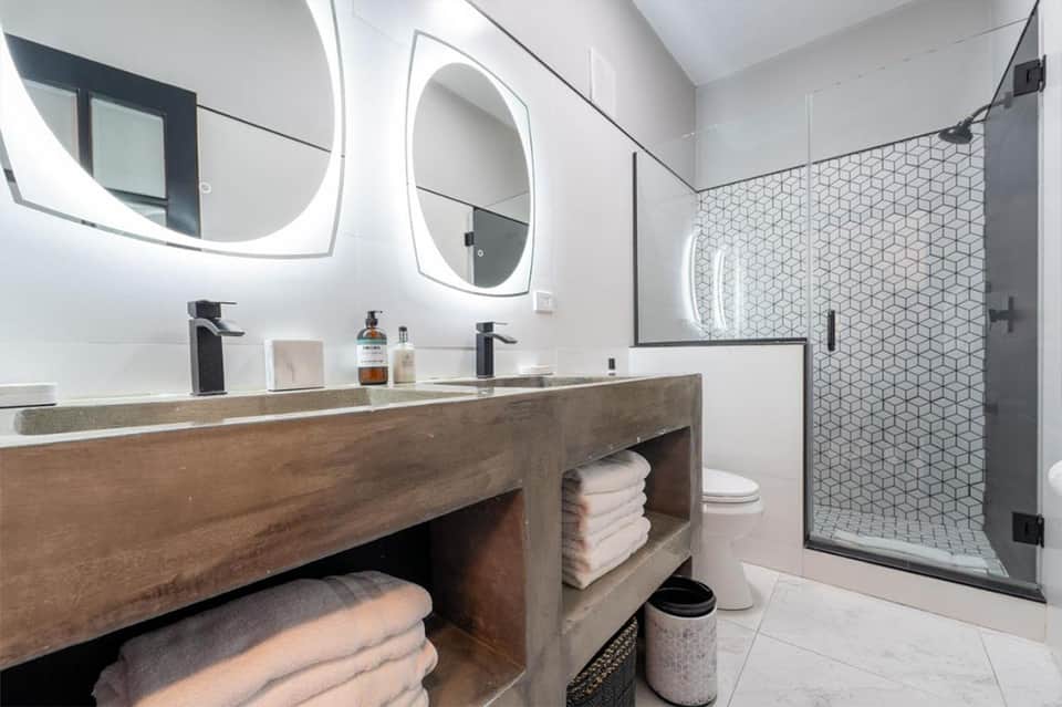 Modern hotel bathroom with dual vanity, LED mirrors, walk-in shower with hexagonal tile, and wood cabinetry