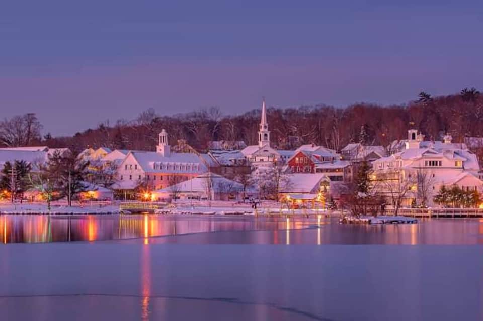 Snowy New England village at dusk with illuminated church steeples reflected in frozen lake