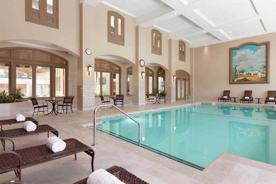 Indoor hotel pool with turquoise water, arched doorways, lounge chairs, towels, and natural skylight ceiling