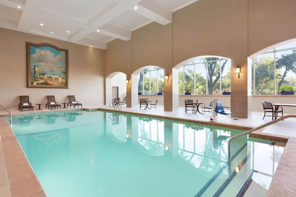 Indoor swimming pool with turquoise water, lounging chairs, arched windows, and tan walls