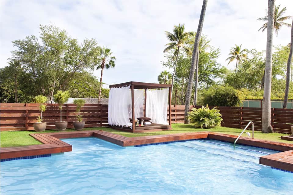 Residential pool with wood decking, brown cabana with white curtains, palm trees, and fenced tropical setting