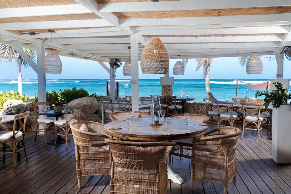 Beachfront dining area with woven pendant lights, wooden tables, ocean view, and palm trees