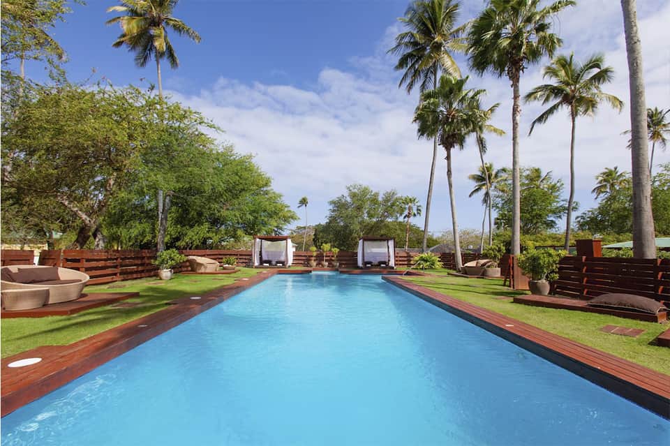 Resort pool surrounded by palm trees, green lawn, wooden deck, and cabana pavilions