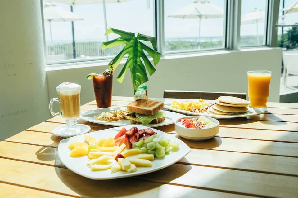 Breakfast spread with fresh fruits, bacon, pancakes, beverages, and potted plant on sunny dining table