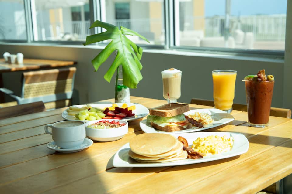 Breakfast spread on wooden table with pancakes, eggs, fresh fruit, beverages, and potted plant by window