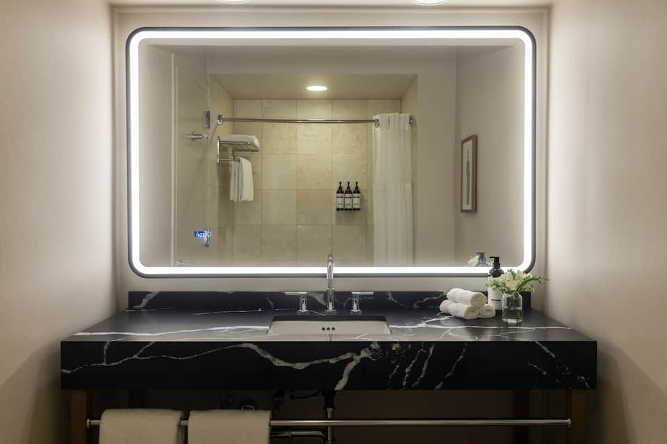 Luxurious bathroom with large illuminated mirror, black marble vanity, glass shower enclosure, and premium toiletries