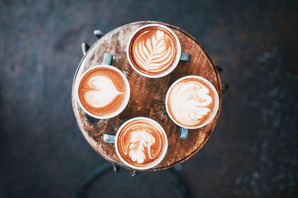 Four latte cups with decorative latte art arranged on a round wooden table, overhead view