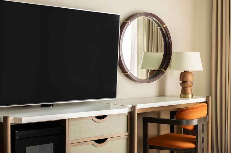 Hotel room desk with flat-screen TV, wooden furniture, round mirror, and table lamp