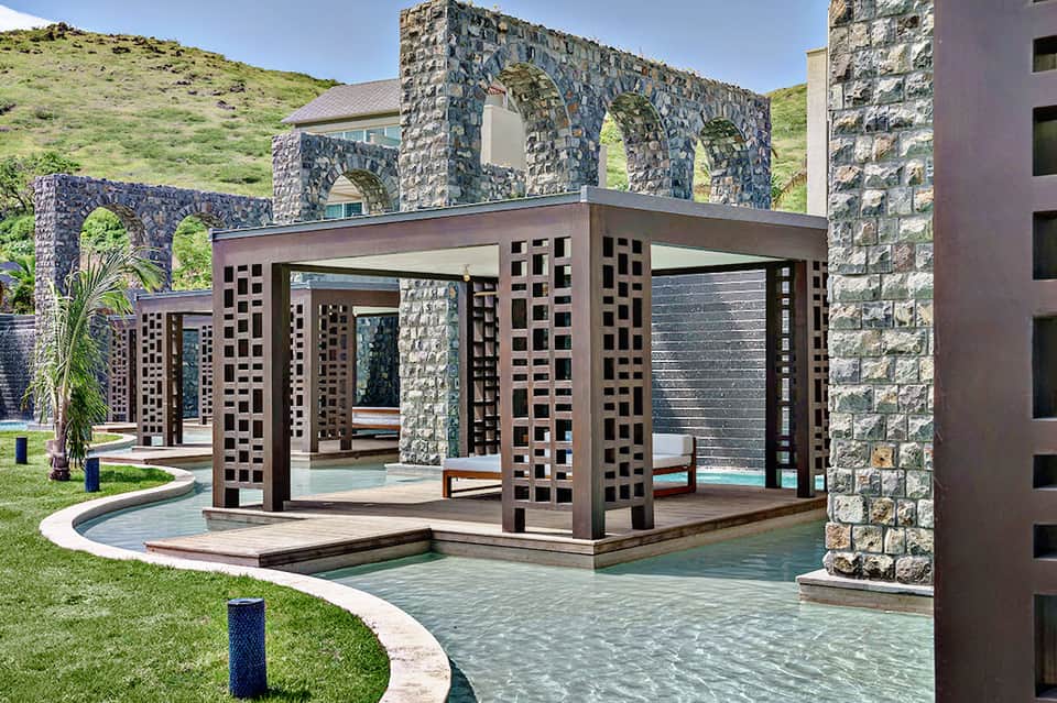 Luxury resort courtyard with stone archways, dark wooden pergola, and cascading water features surrounded by green hillside