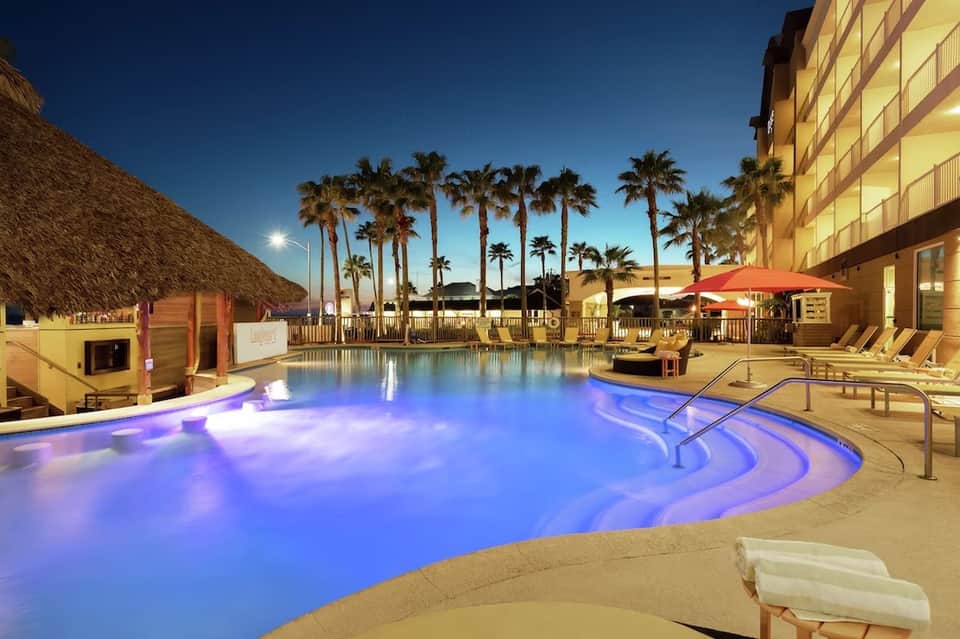 Illuminated resort pool with purple ambient lighting at night, palm trees, and tiki hut in background