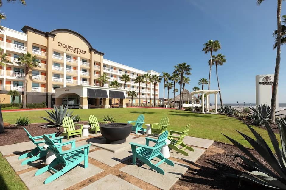 Beachfront resort with palm trees, colorful Adirondack chairs around fire pit, and ocean view in background