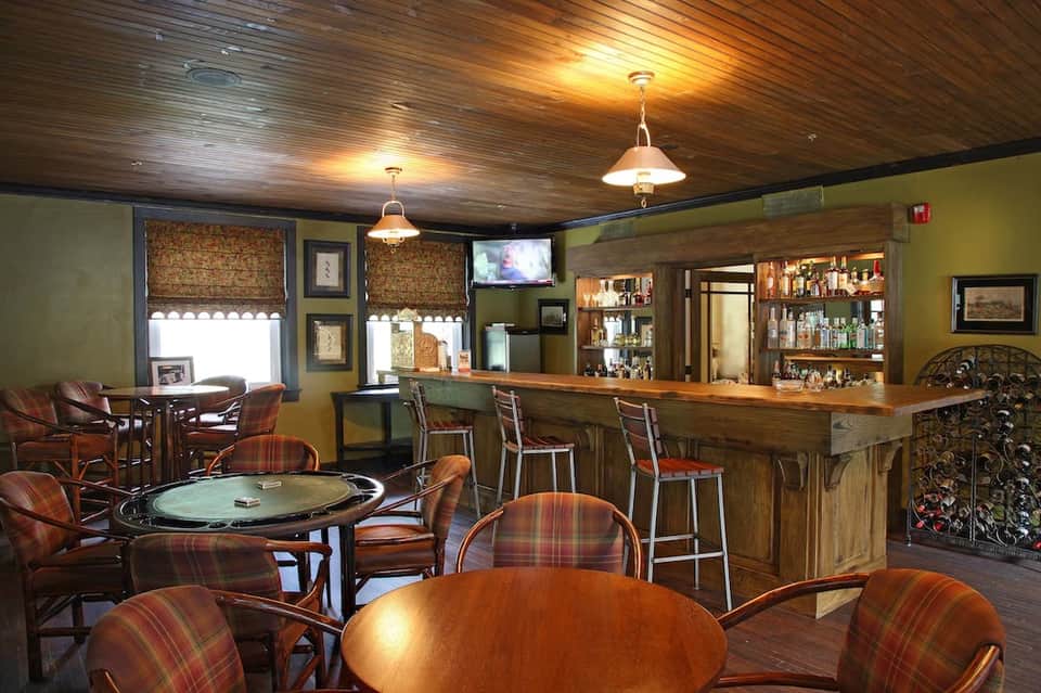 Casual bar lounge with wood ceiling, green walls, plaid upholstered chairs, tables, and full liquor bar