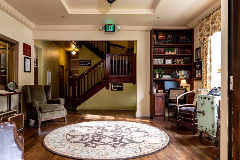 Hotel lobby entrance with wooden staircase, armchair, bookshelves, round rug, and vintage decor