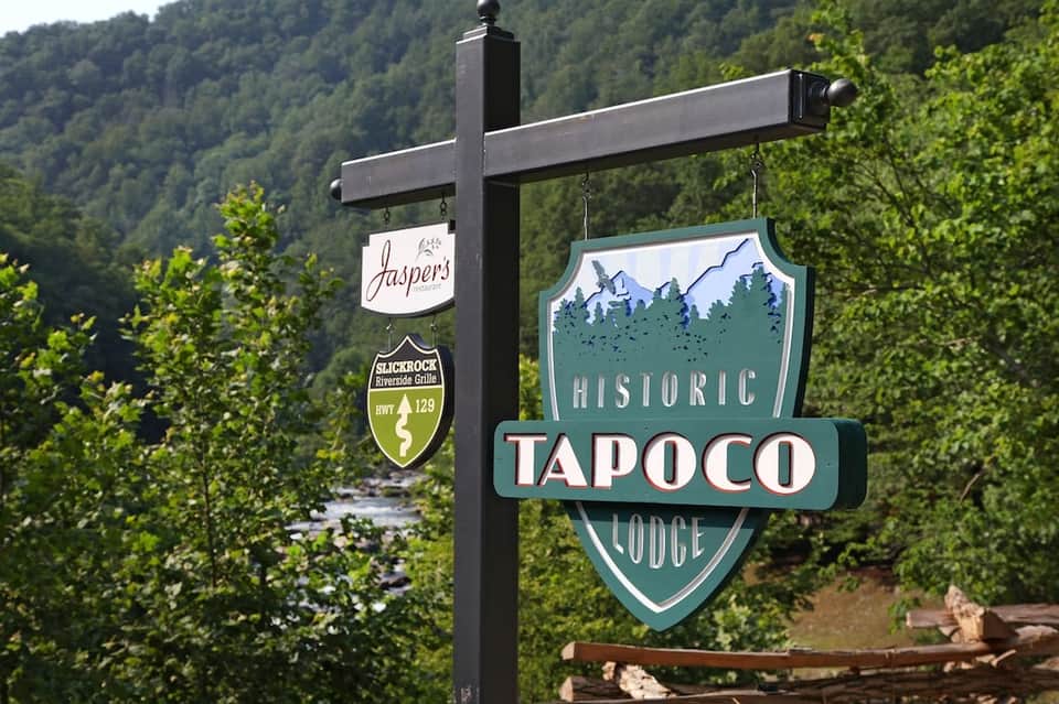 Historic Tapoco Lodge sign with Jaspers restaurant sign hanging on black post in forested mountain valley setting