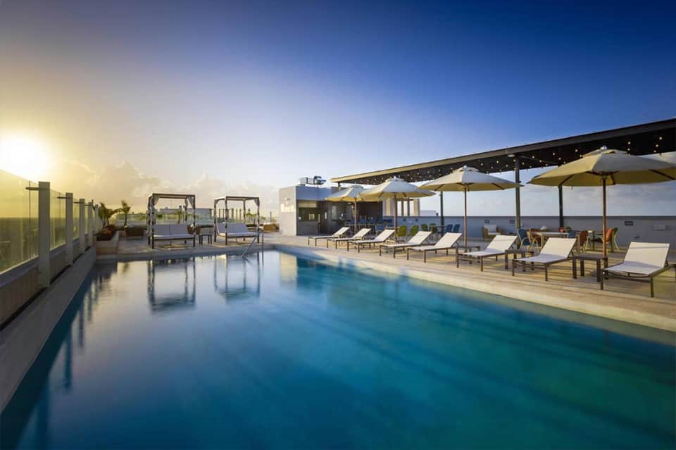 Modern rooftop infinity pool at sunset with yellow umbrellas, lounge chairs, and ocean horizon