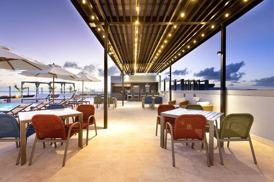Modern rooftop terrace at dusk with dining tables, colorful chairs, and pool in background
