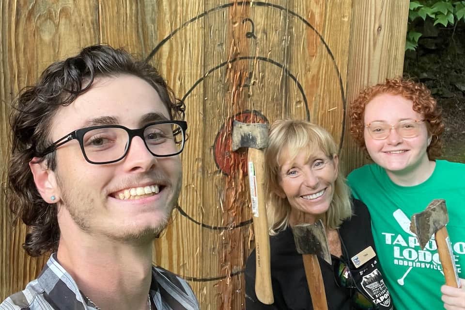 Three people posing indoors with axes and rustic wooden wall backdrop