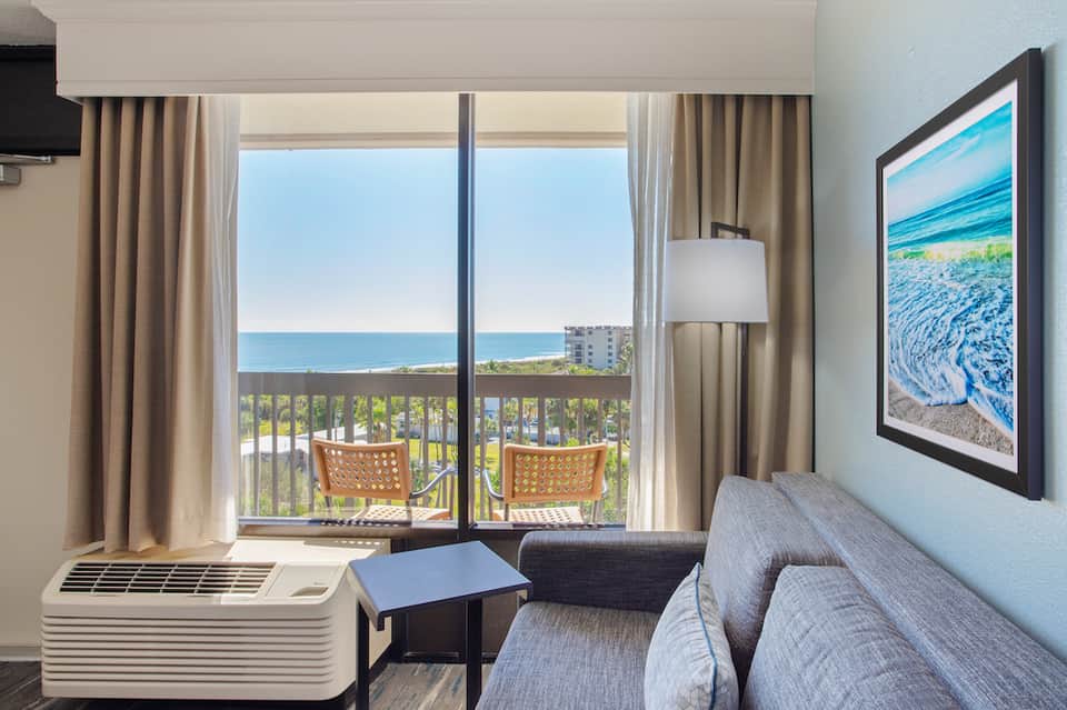 Hotel room with ocean view balcony, beige curtains, seating area, and framed beach artwork on light blue wall.