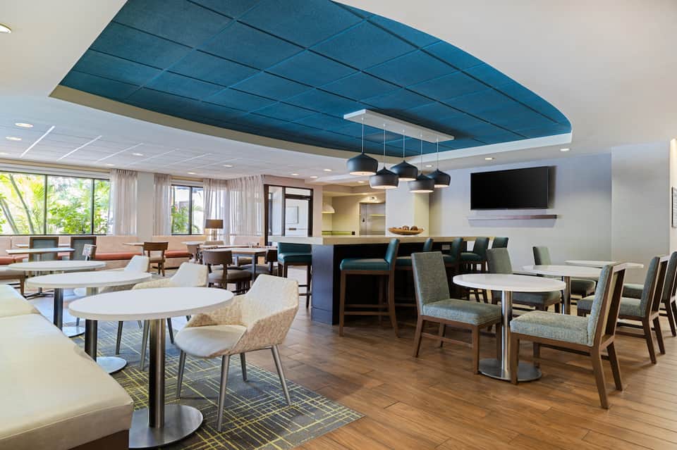 Hotel common area with round white tables, modern chairs, blue ceiling accent, and large windows