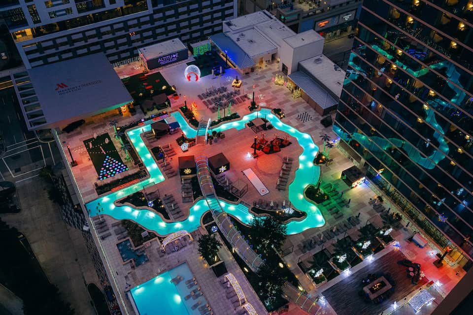 Aerial view of illuminated resort courtyard with glowing turquoise pool and surrounding buildings at night