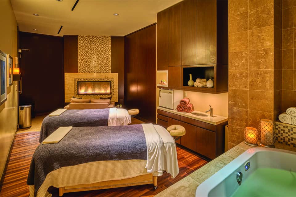 Luxury spa treatment room with fireplace, two massage beds, and bronze tile accents